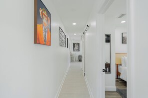 Um corredor com obras de arte nas paredes, uma porta que dá para um quarto e uma pequena mesa com abajur.