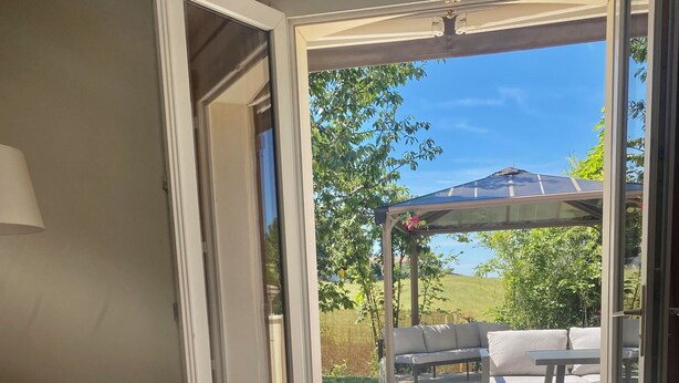 Eine geöffnete Schiebetür führt zu einer Außenpatio-Terrasse mit überdachter Sitzgruppe und Blick auf eine grüne Landschaft.