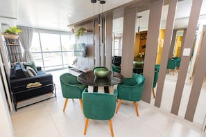 Ein modernes Wohnzimmer mit einem schwarzen Sofa, grünen Sesseln, einem runden Tisch mit einer grünen Schale und einem Fernseher an der Wand.