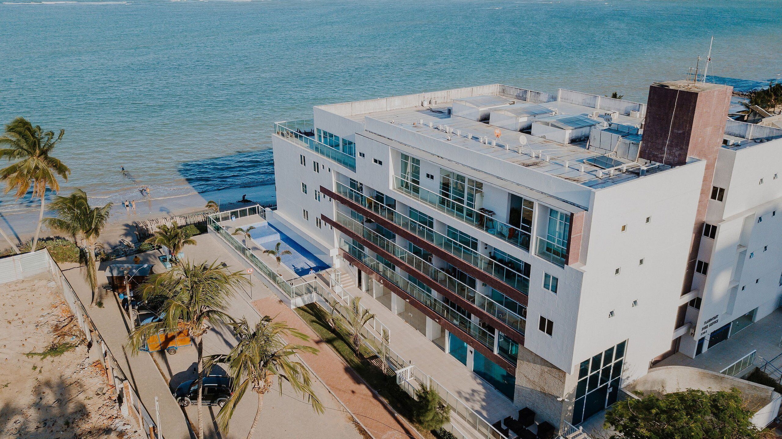 Un edificio moderno de varios pisos con amplias ventanas, una terraza en la azotea y vista al mar.