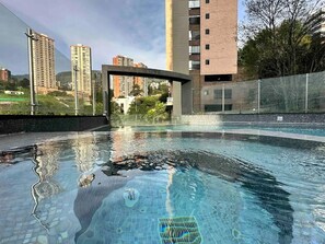 Una piscina con agua cristalina, rodeada por una valla y edificios al fondo.