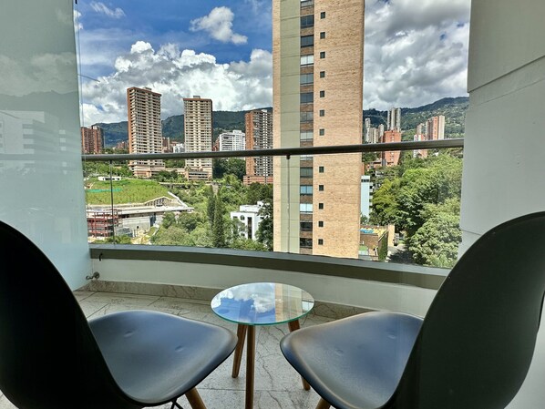 Un balcón con dos sillas y una mesita pequeña, con vistas a un paisaje urbano con rascacielos y zonas verdes.