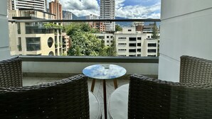 Ein Balkon mit Korbmöbeln und einem Glastisch, mit Blick auf eine Stadtlandschaft aus Gebäuden und Bäumen.