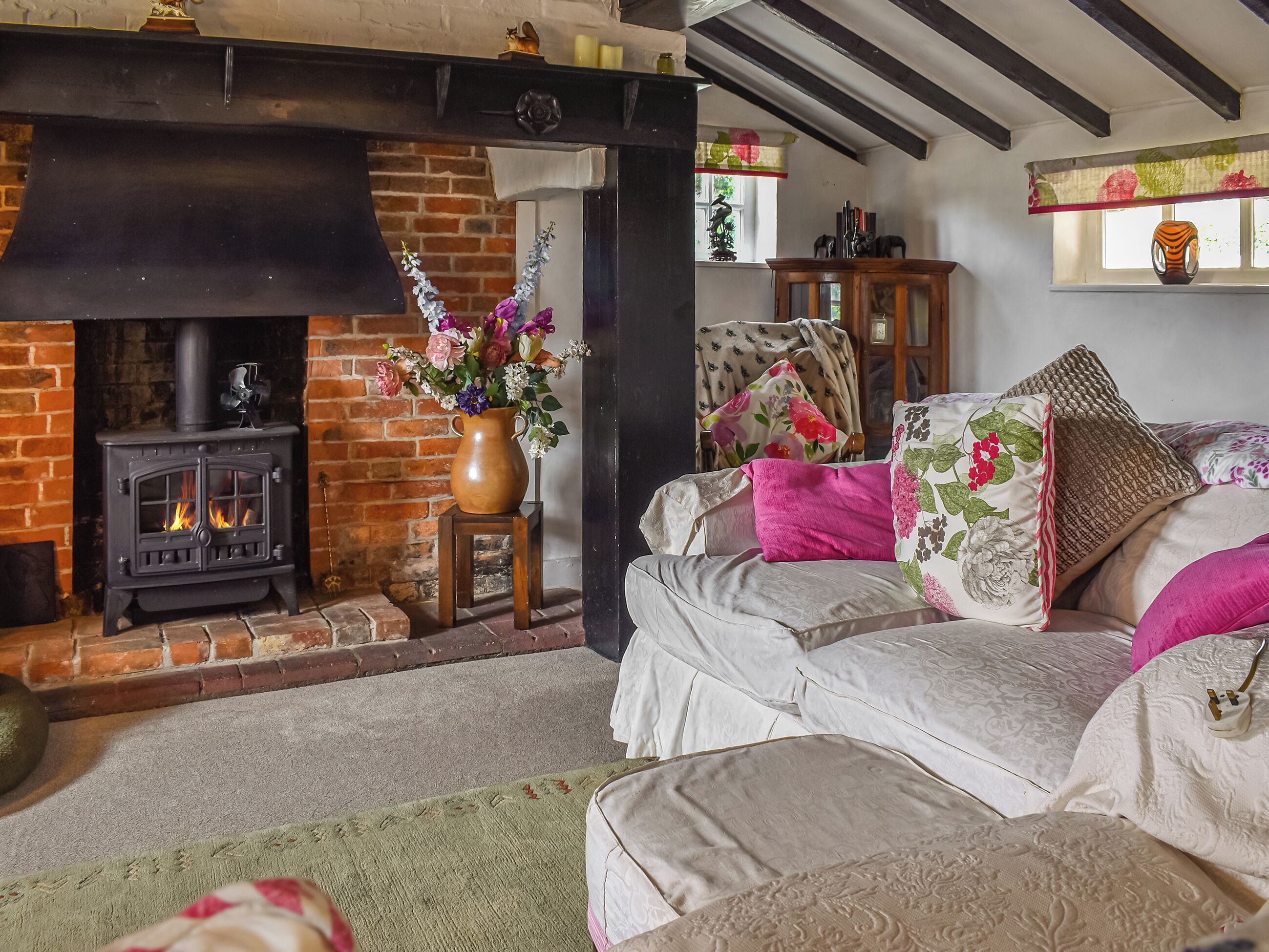 A cozy living room with a fireplace, a sofa with floral cushions, and a vase of flowers.