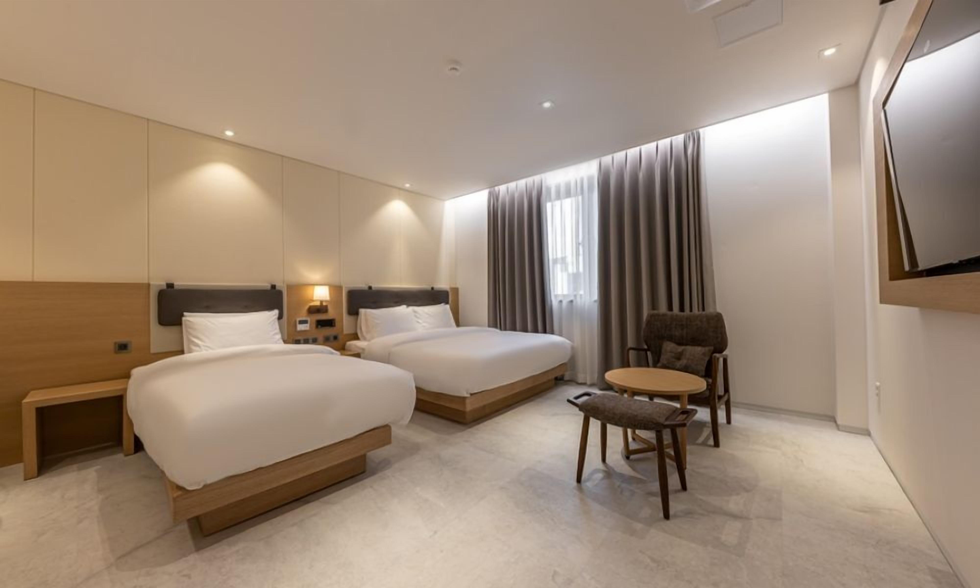 A modern hotel room with two beds, a small round table, a chair, and a TV mounted on the wall.