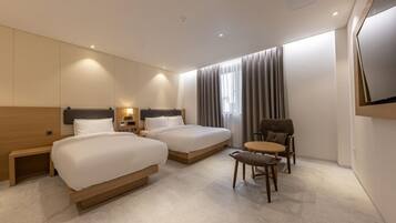A modern hotel room with two beds, a small round table, a chair, and a TV mounted on the wall.