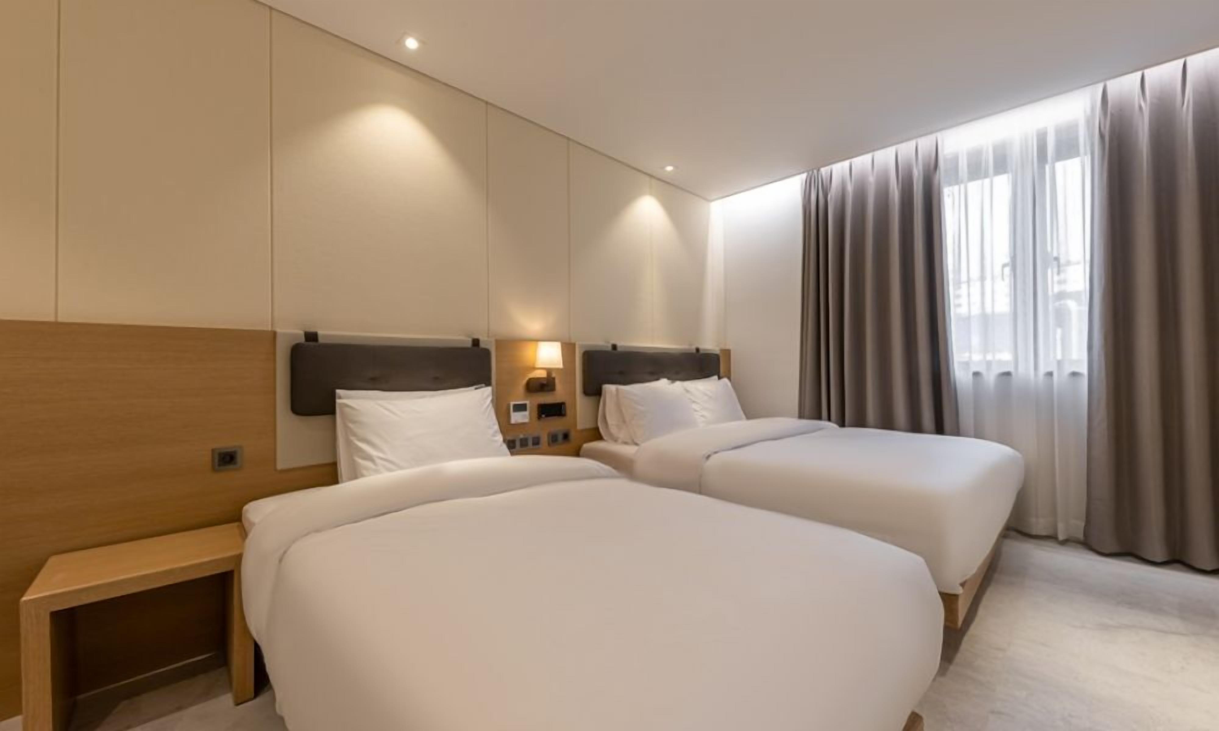 A hotel room with two beds, wooden headboards, a bedside table, a lamp, and a window with curtains.