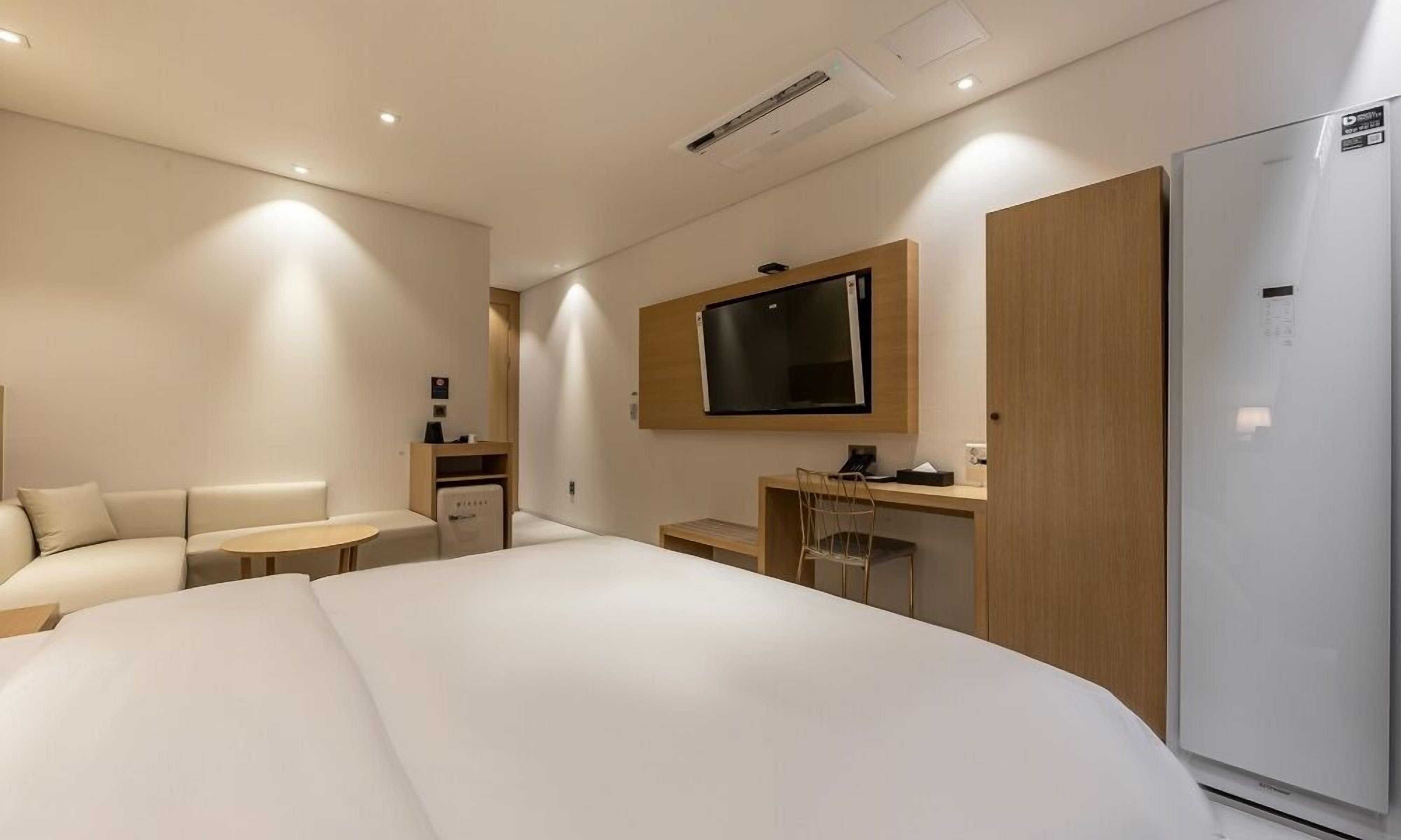 A modern hotel room with a large bed, a desk with a chair, a television mounted on the wall, and a refrigerator.
