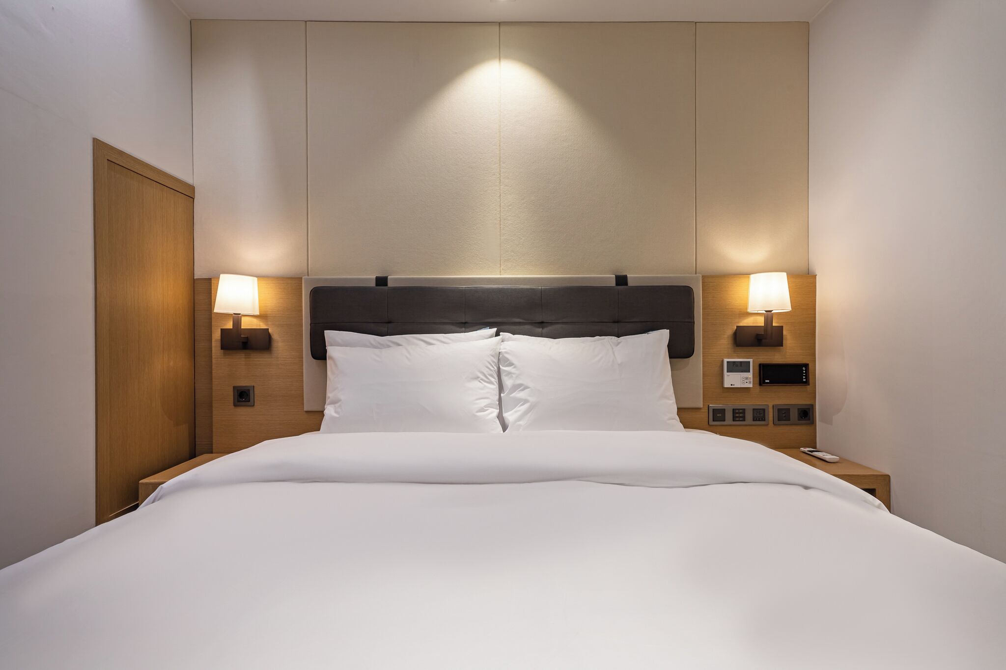 Een netjes opgemaakt bed met een houten hoofdeinde, twee bedlampen en een klein nachtkastje met een telefoon en een afstandsbediening.