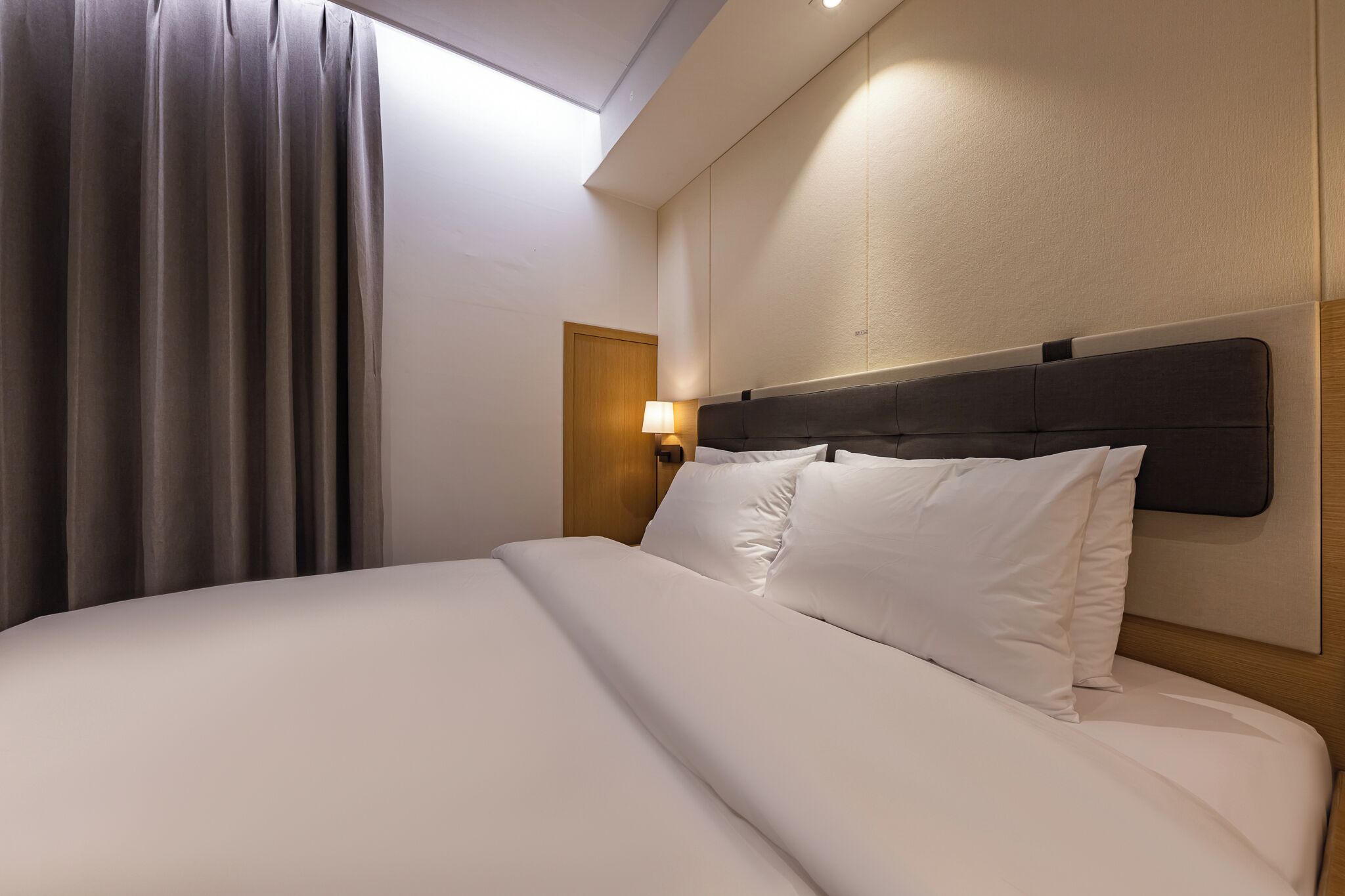 A neatly made bed with white linens, a dark headboard, and a bedside lamp.