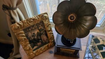 A vintage gramophone on a wooden table with a framed picture beside it, near a window with a view of trees.