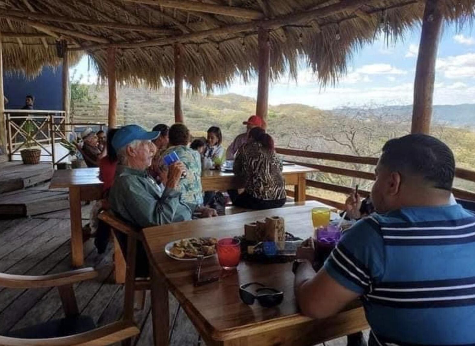 A group of people dining at an outdoor wooden table with a thatched roof, overlooking a mountainous landscape.