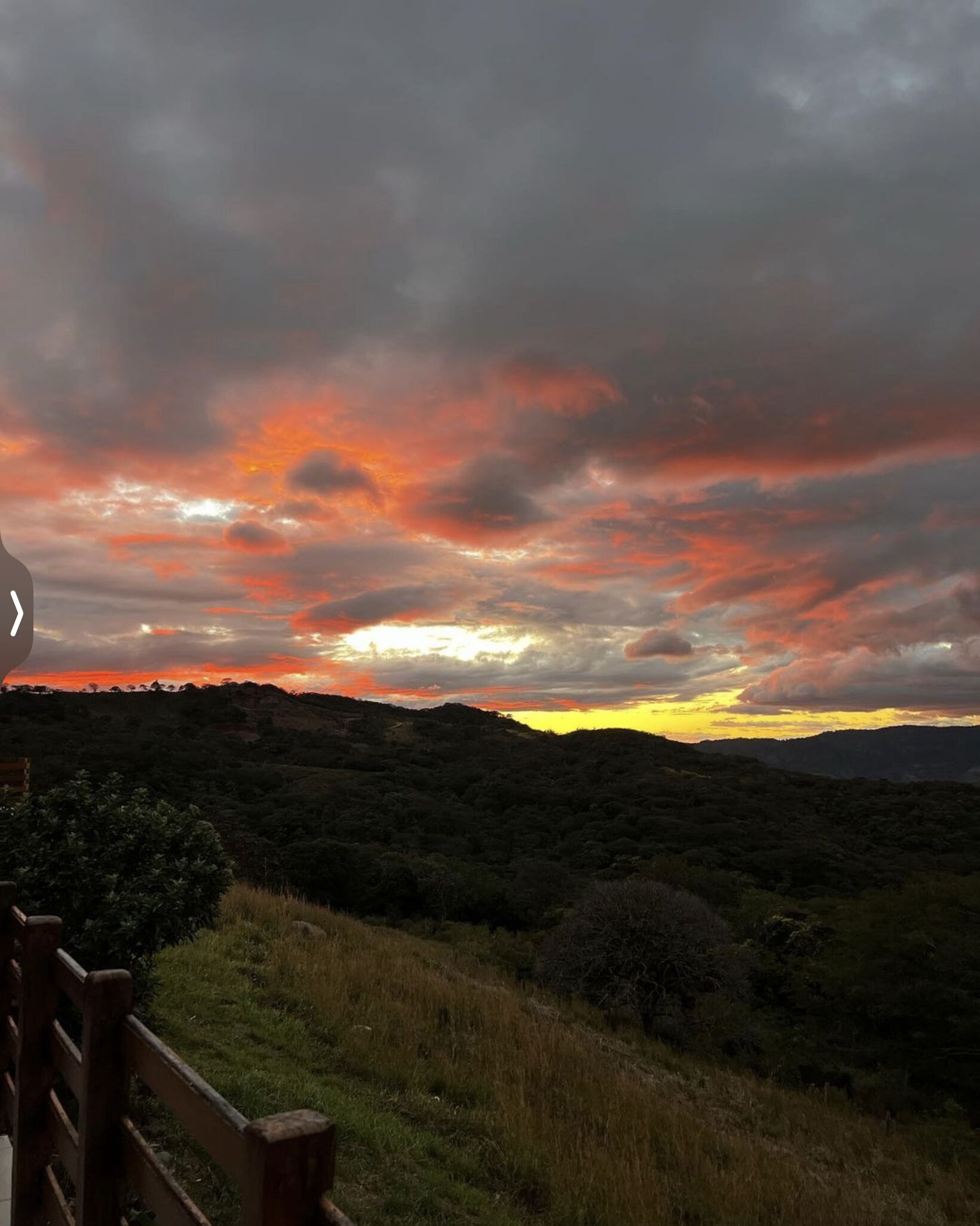 A landscape with a sunset over a hilly area, a wooden fence in the foreground, and a view of the sky with clouds.