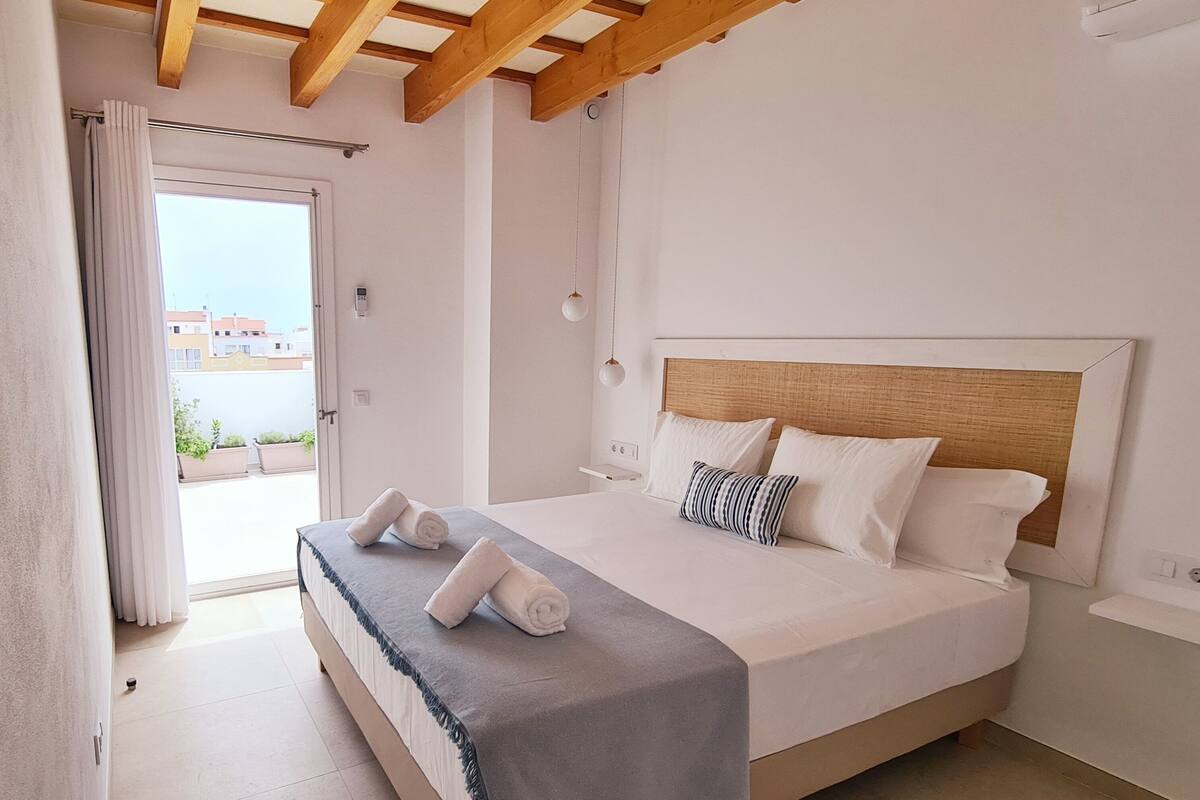 Un dormitorio con cama, techo de madera, ventana con cortinas y vistas a edificios.