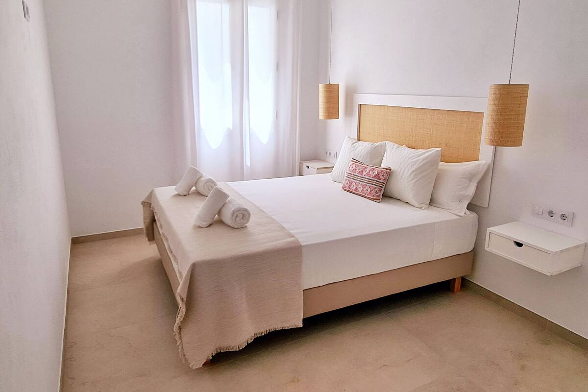 Una cama bien hecha con ropa blanca y un cojín estampado, una mesita de noche con una lámpara, y una ventana con cortinas traslúcidas.