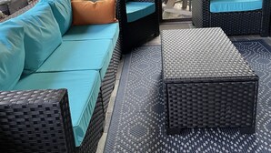 Un patio couvert aménagé avec des meubles en osier, notamment un canapé et deux poufs, et un tapis à motifs sur le sol.