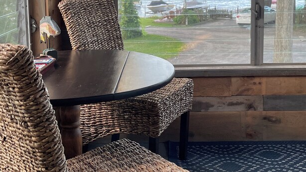 L’intérieur d’un chalet en bois, avec une table ronde et des chaises en osier, une grande fenêtre donnant sur un lac où l’on aperçoit des bateaux et un quai.