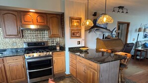 Une cuisine avec des meubles de rangement en bois, une cuisinière à gaz et un grand îlot central doté d’un évier. Des luminaires sont suspendus au-dessus de l’îlot, et un coin repas comprend une table et des chaises.