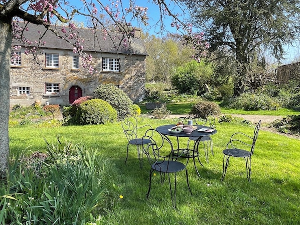 A stone building with a red door, surrounded by a well-maintained garden with a table and chairs. - Farmhouse and Cottage with heated pool, Sleeps 9 (Saint-Nicolas-du-Pélem)