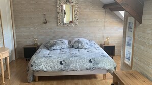 A bedroom with a wooden floor, a bed with a floral bedspread, a wooden bench, a wall-mounted mirror, and a wooden ceiling with exposed beams. - Loft 1er Étage (Andernos-les-Bains)