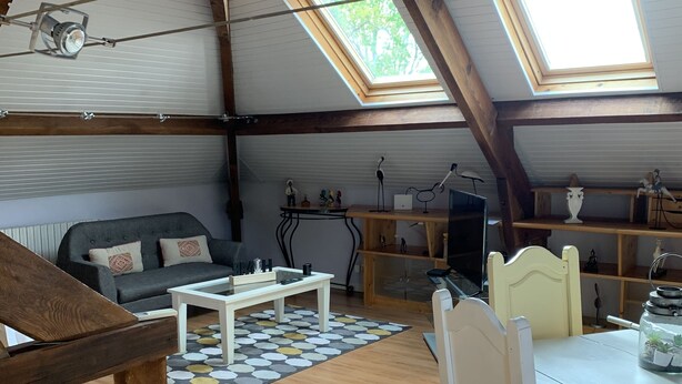 A cozy attic room with a sloped ceiling, wooden beams, a skylight, a sofa, a dining table, and a small kitchen area. - Loft 1er Étage (Andernos-les-Bains)