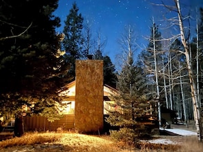 Une cabane dotée d’une cheminée en pierre, entourée d’arbres et sous un ciel nocturne dégagé.