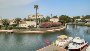 Uma marina com barcos atracados, um canal navegável e edifícios com palmeiras e arbustos floridos.