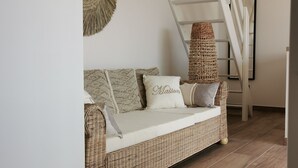 Um sofá de vime com uma almofada branca e uma almofada com a inscrição "Maison", em frente a uma parede branca, com uma cesta trançada no chão e uma escada branca ao fundo.