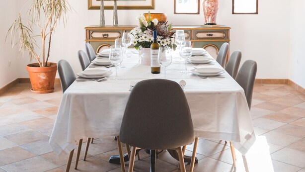Un coin repas avec une longue table dressée pour six personnes, entourée de chaises aux pieds en bois. Un pot de fleurs se trouve à gauche, et une lampe à droite.
