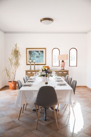 Een eethoek met een lange tafel gedekt voor zes personen, omringd door stoelen met houten poten. Links staat een potplant en rechts een lamp.