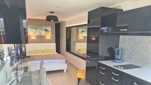 Moderni hotellihuone, jossa on lasinen työpöytä, keltainen koristetyynyillä varustettu sänky, pieni keittiö, jossa on kahvinkeitin, sekä taideteoksia seinillä.