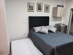A bedroom with a bed, a nightstand, a trash bin, and three framed pictures on the wall. - Lindo Apartamento en Laureles Medellín (Medellín)