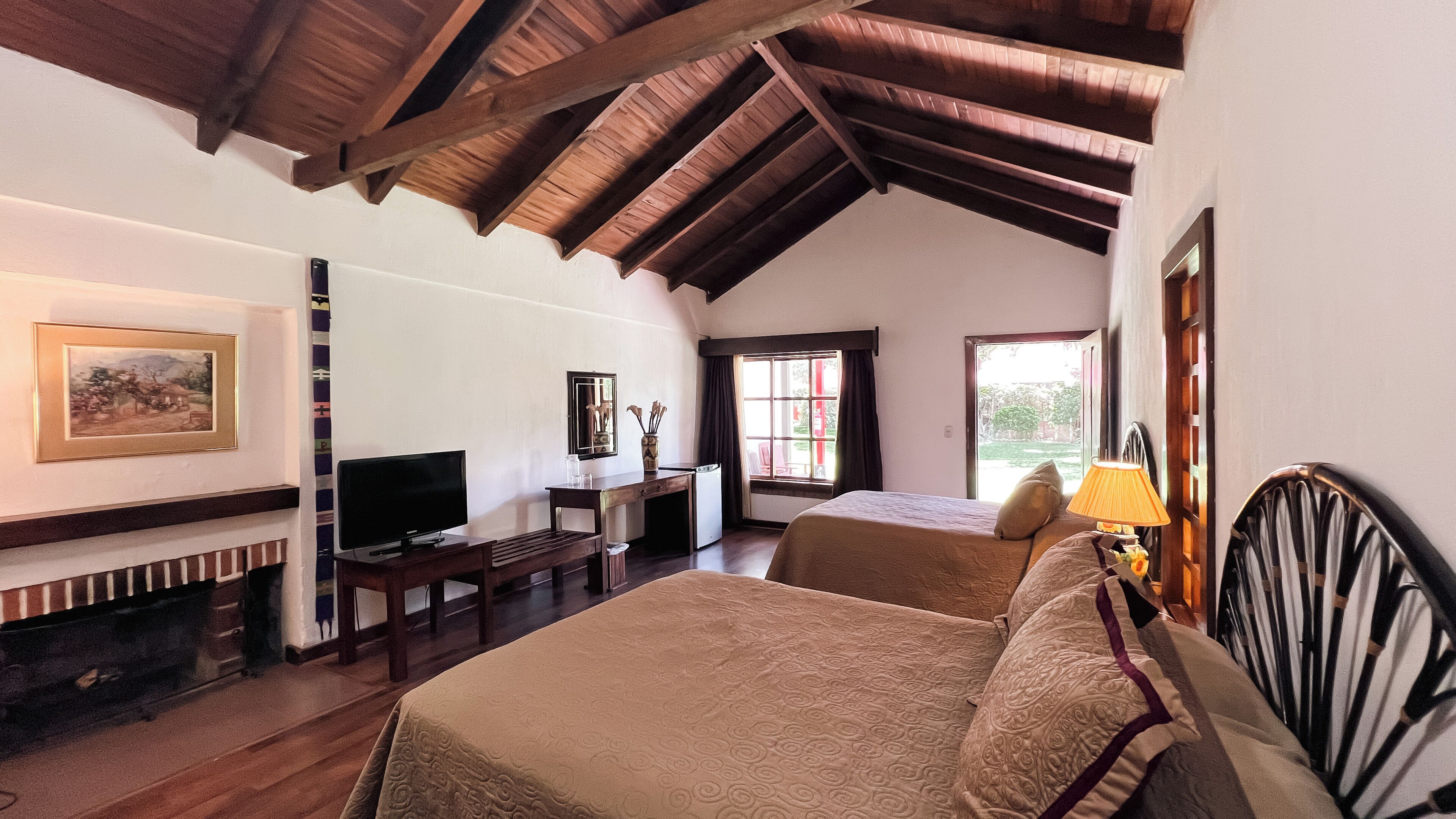 Un dormitorio con techo de madera, una cama, un escritorio con televisor y un ventanal con cortinas.