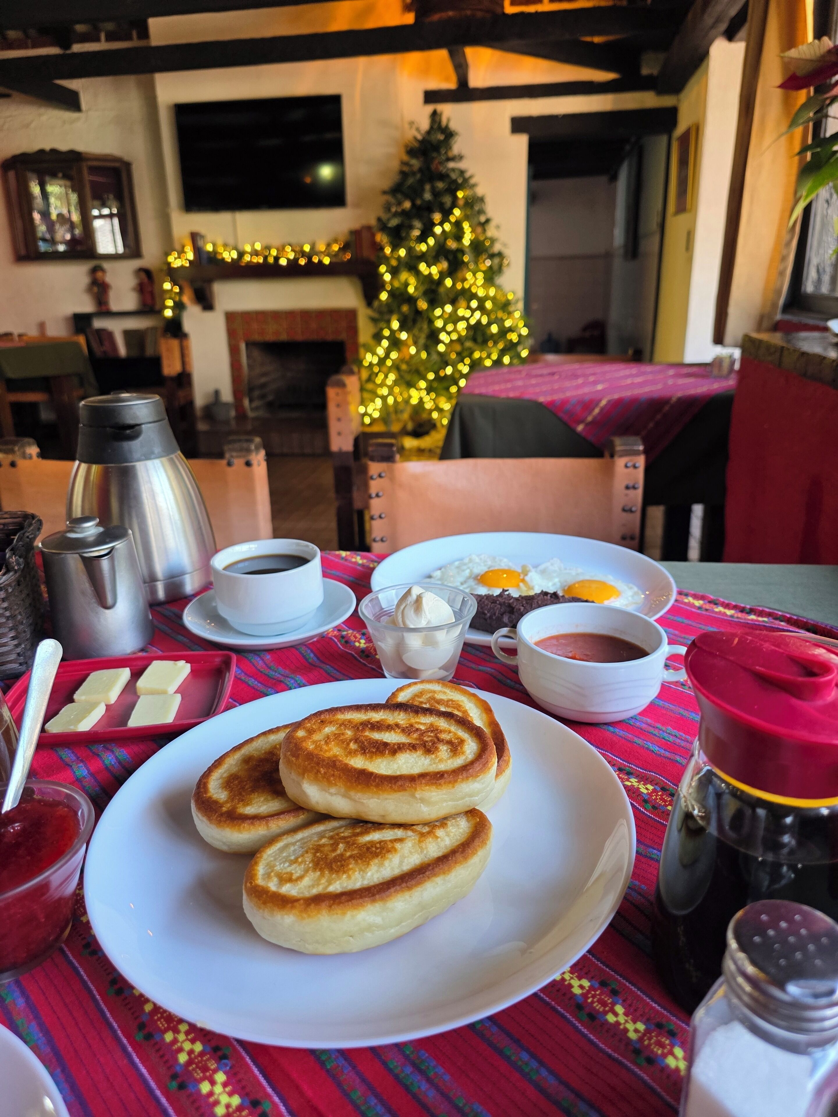 Une table à manger dressée avec des aliments pour le petit-déjeuner, notamment des crêpes et des œufs, et un sapin de Noël en arrière-plan.