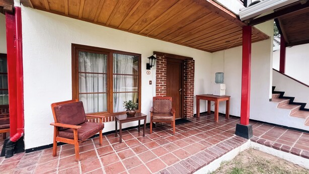 A covered patio area with a wooden ceiling, a brick wall, and a tiled floor. There are two wooden chairs, a small table with a potted plant, and a water dispenser on a table. - Hotel Rancho Grande (Panajachel)