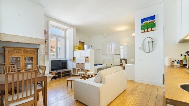 A modern living room with a sofa, wooden dining table, and chairs. There is a kitchen area with cabinets and a window. - Fully new refurbished Alfama Central Cosy Home (Lisboa)