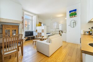 A modern living room with a sofa, wooden dining table, and chairs. There is a kitchen area with cabinets and a window. - Fully new refurbished Alfama Central Cosy Home (Lisboa)