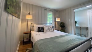Um quarto com uma cama, duas luminárias de cabeceira, um criado-mudo, uma planta em vaso e um quadro na parede.