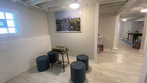 Um quarto com teto branco, janela com persianas, quadro pendurado na parede, mesa pequena com um jogo de tabuleiro e dois pufes.