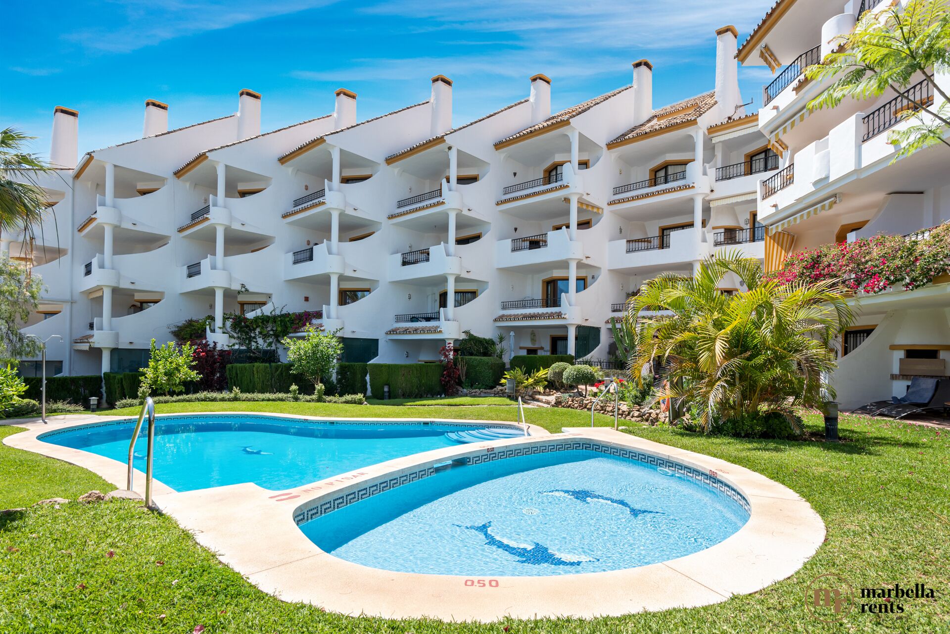 Un hotel de varios pisos con zona de piscina, con un diseño moderno, balcones blancos y un cielo azul despejado.