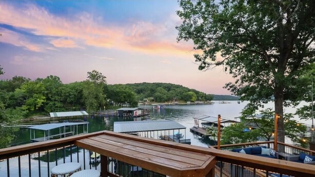 Une terrasse en bois, agrémentée de chaises en osier, qui offre une vue sur un lac, entouré de verdure et bordé de bateaux amarrés.