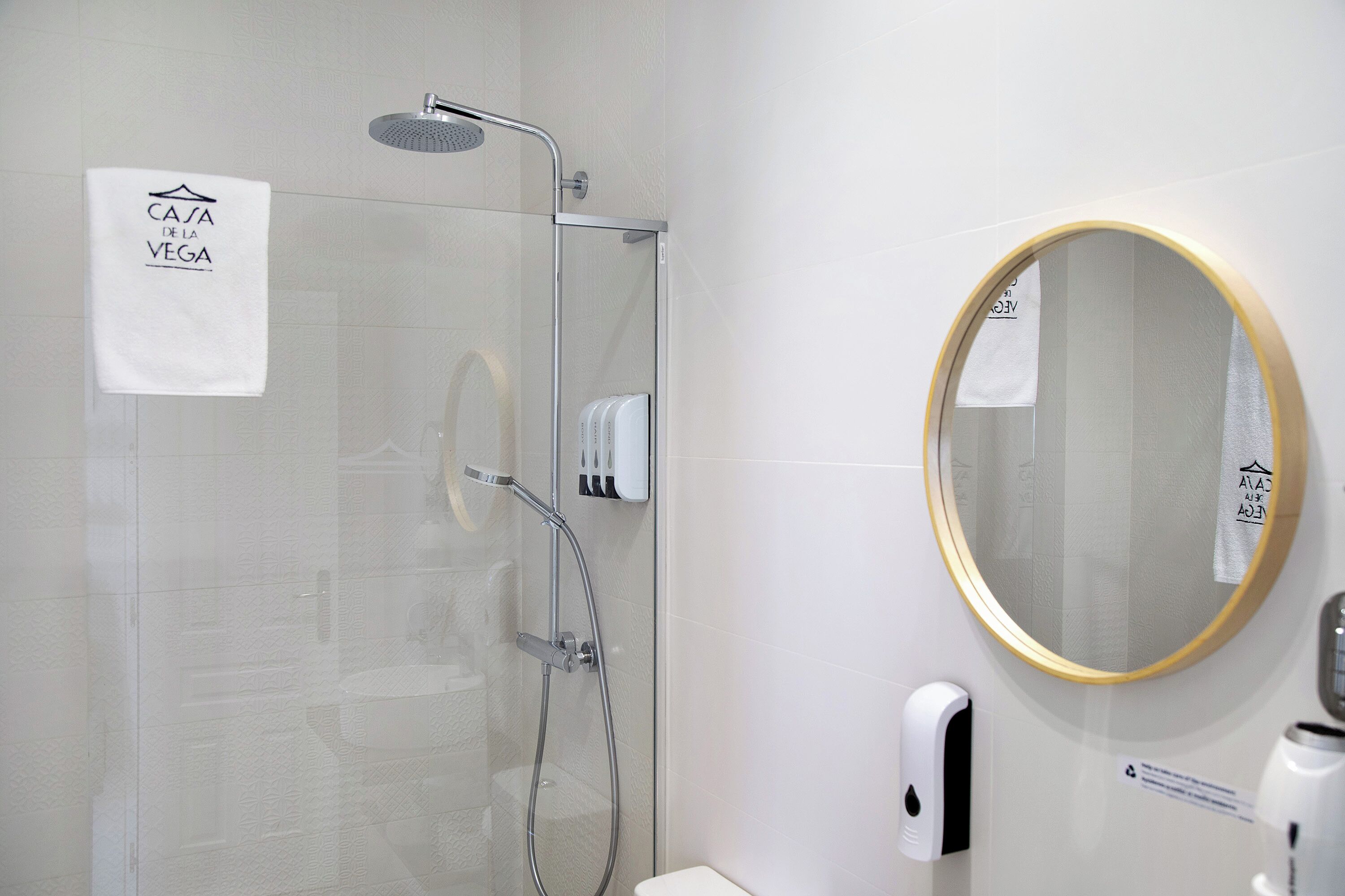 A modern bathroom with a glass-enclosed shower, a round mirror with a gold frame, and a towel hanging on the wall.
