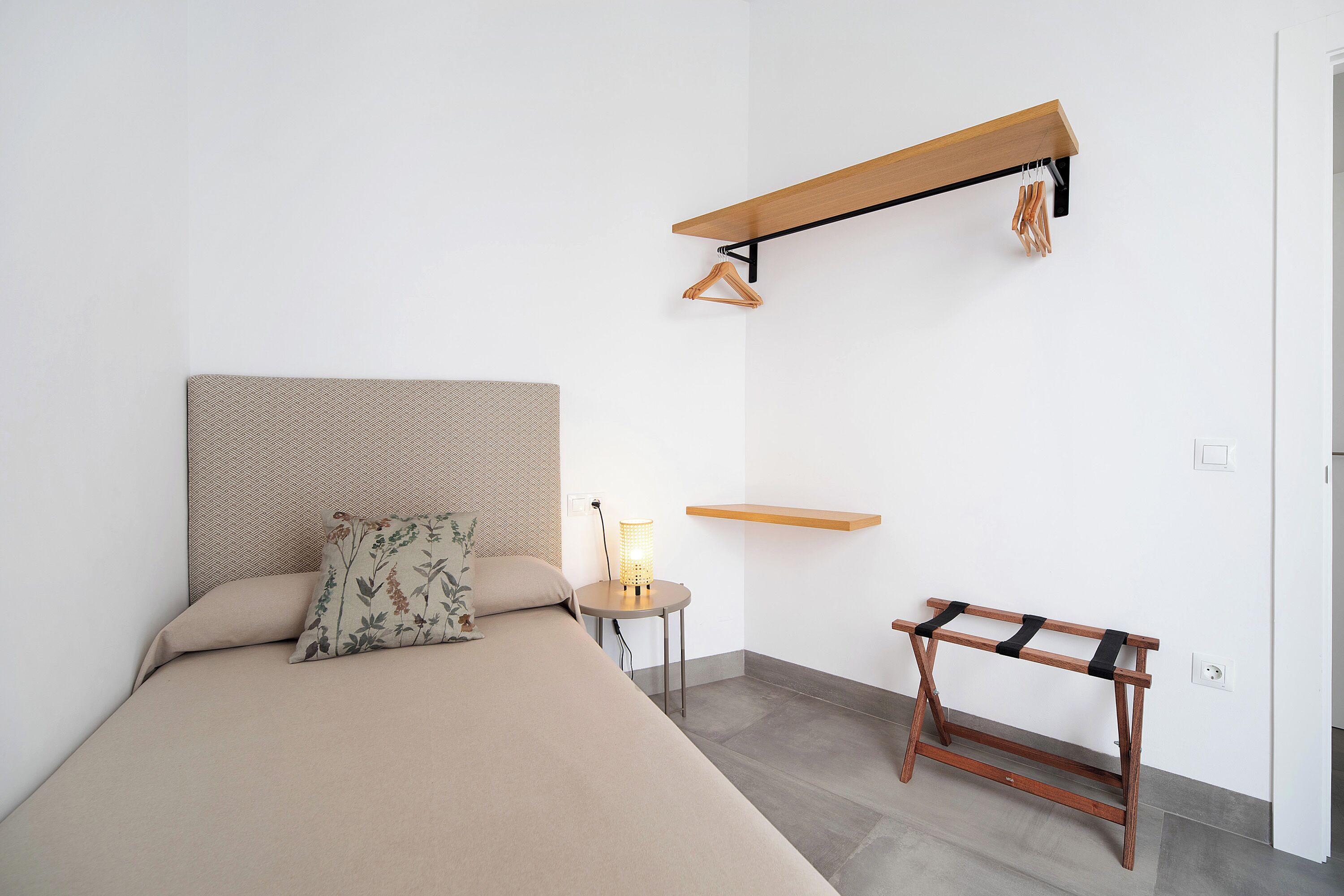 Un dormitorio con una cama, una mesita de noche con lámpara, un estante de madera y un pequeño banco de madera.