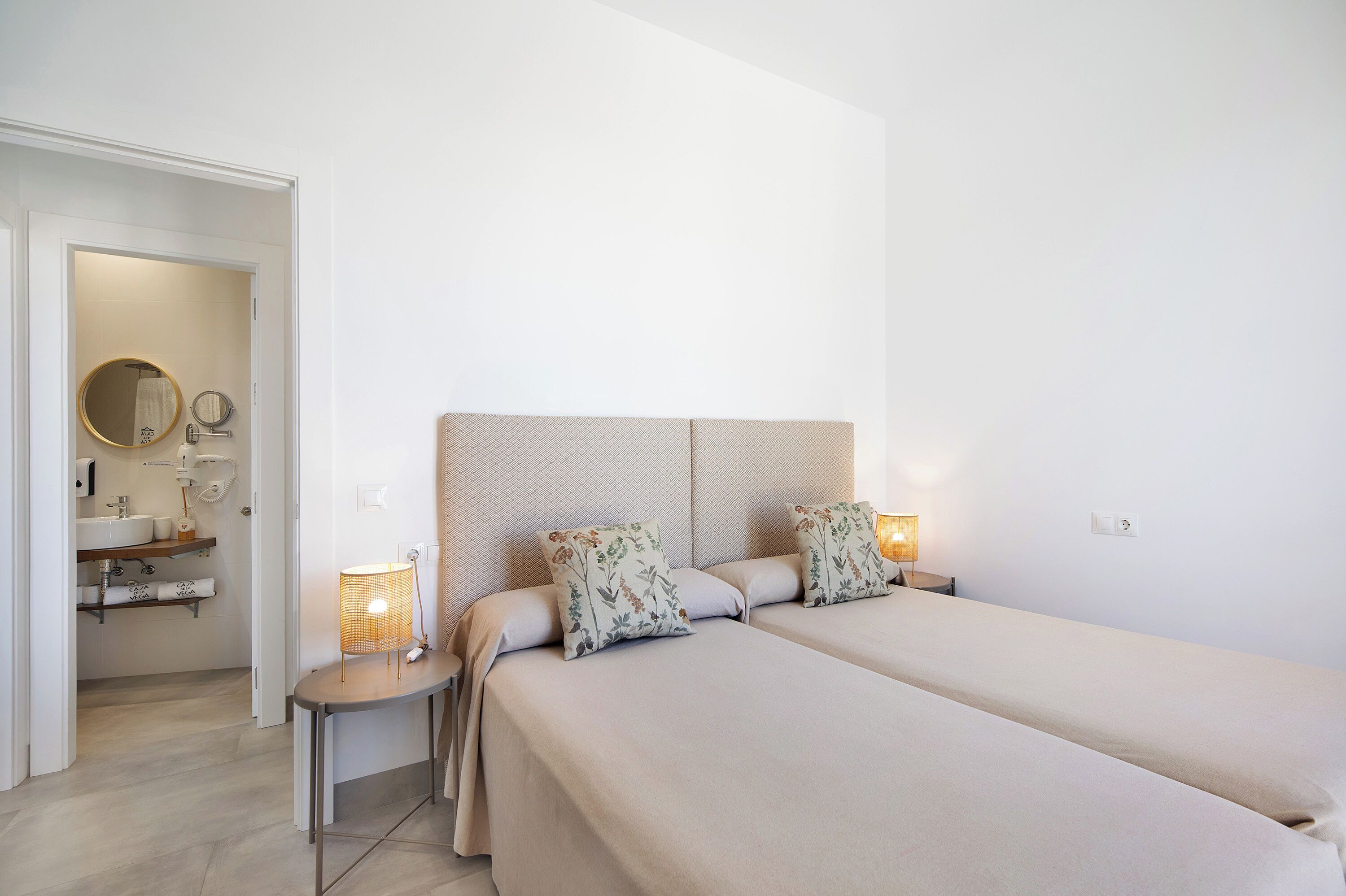 A hotel room with a bed, bedside tables, a nightstand, a mirror, and a bathroom visible through an open door.