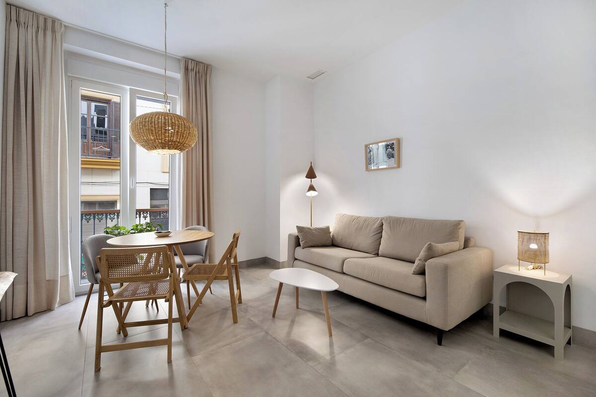 A modern living room with a sofa, a dining table with chairs, a hanging light fixture, and a small side table.