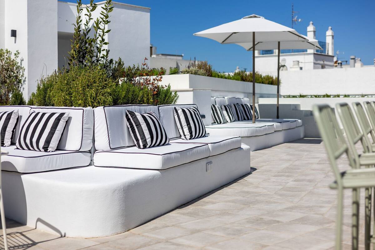 A modern outdoor seating area with white cushioned sofas and striped pillows, surrounded by greenery and a clear blue sky.