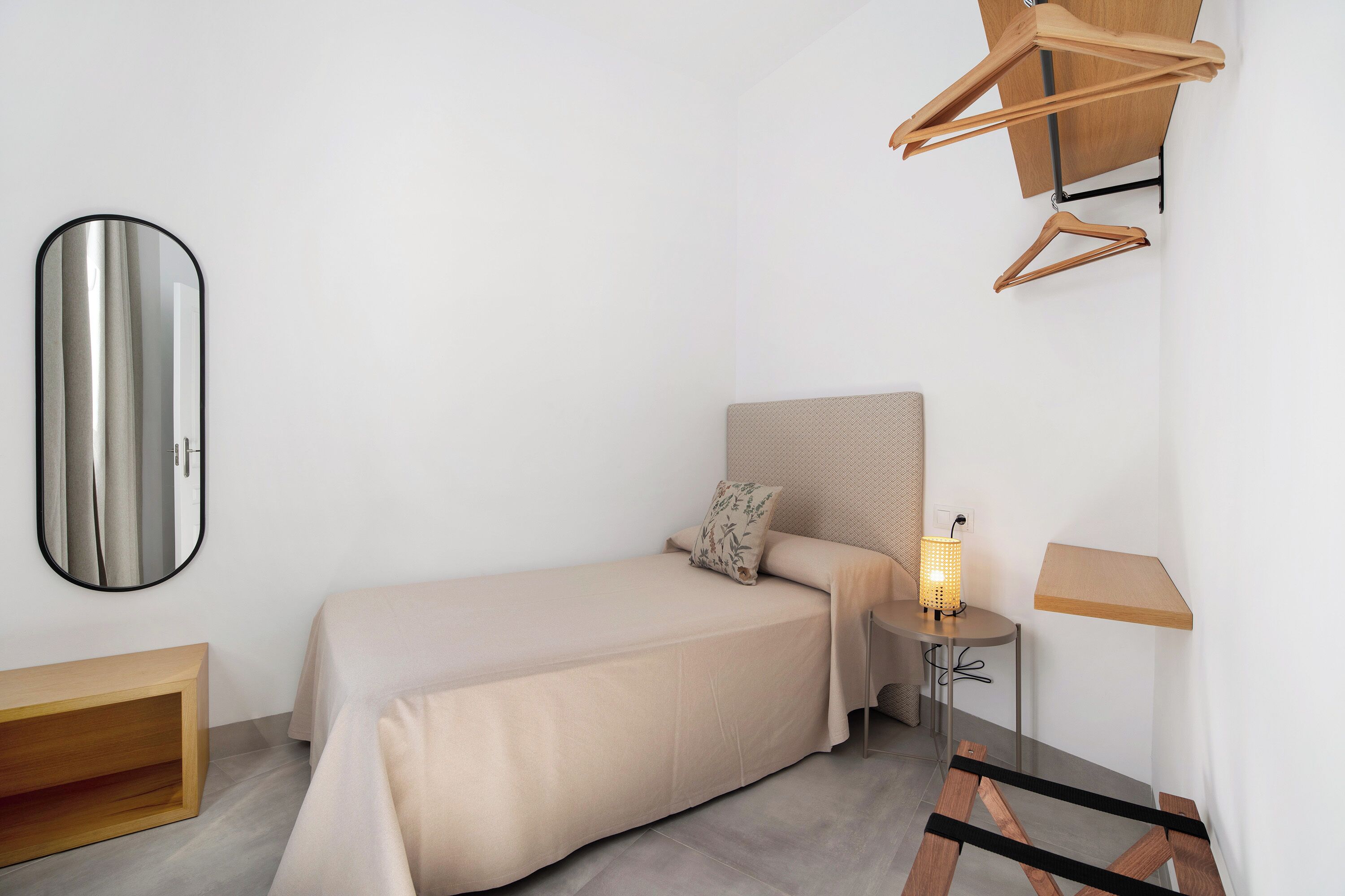 A minimalist bedroom with a bed, bedside table, lamp, and a mirror.