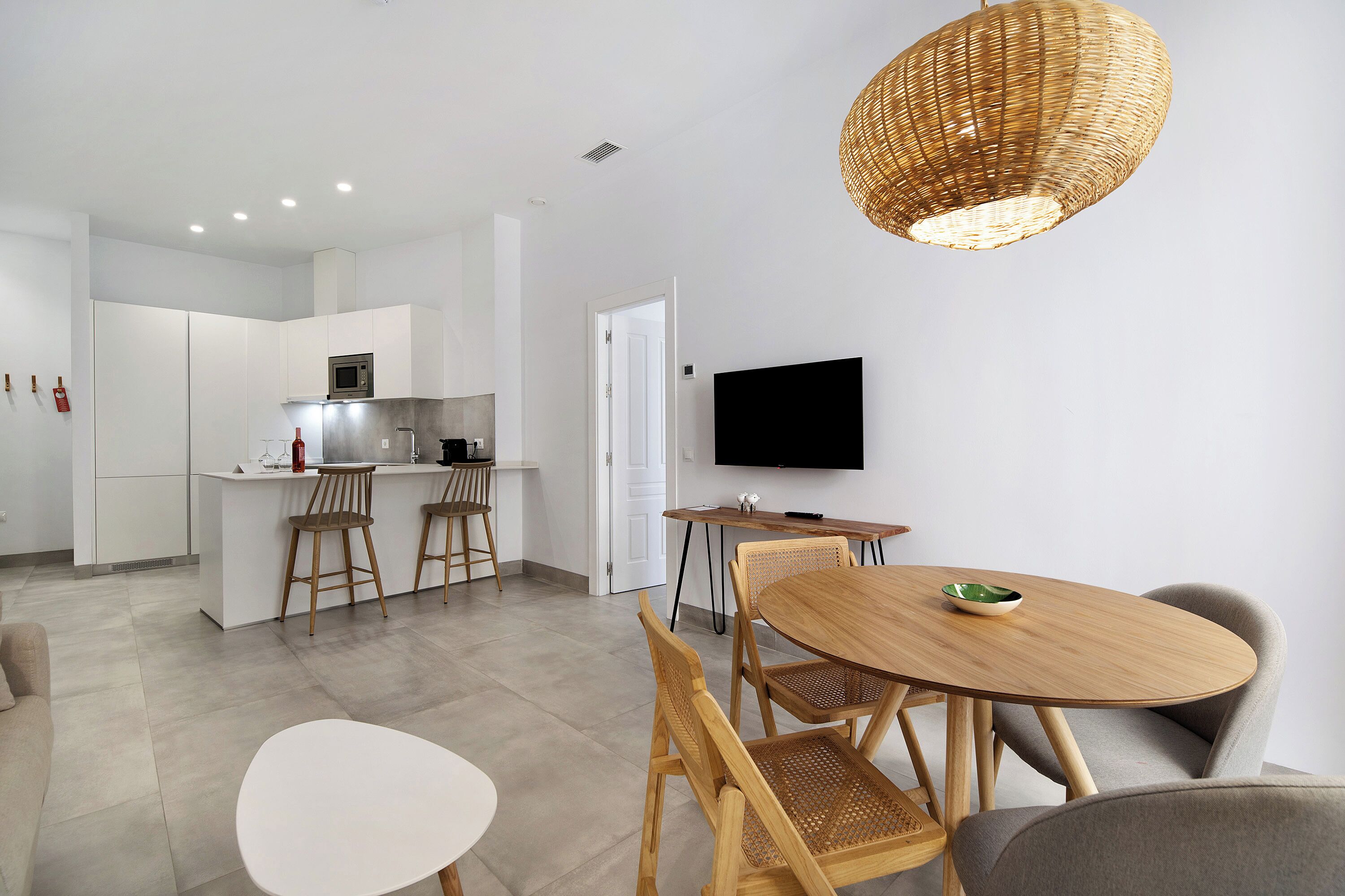Une cuisine moderne avec des meubles blancs, un coin repas doté d’une table ronde en bois et de chaises, et un grand téléviseur à écran plat fixé au mur.
