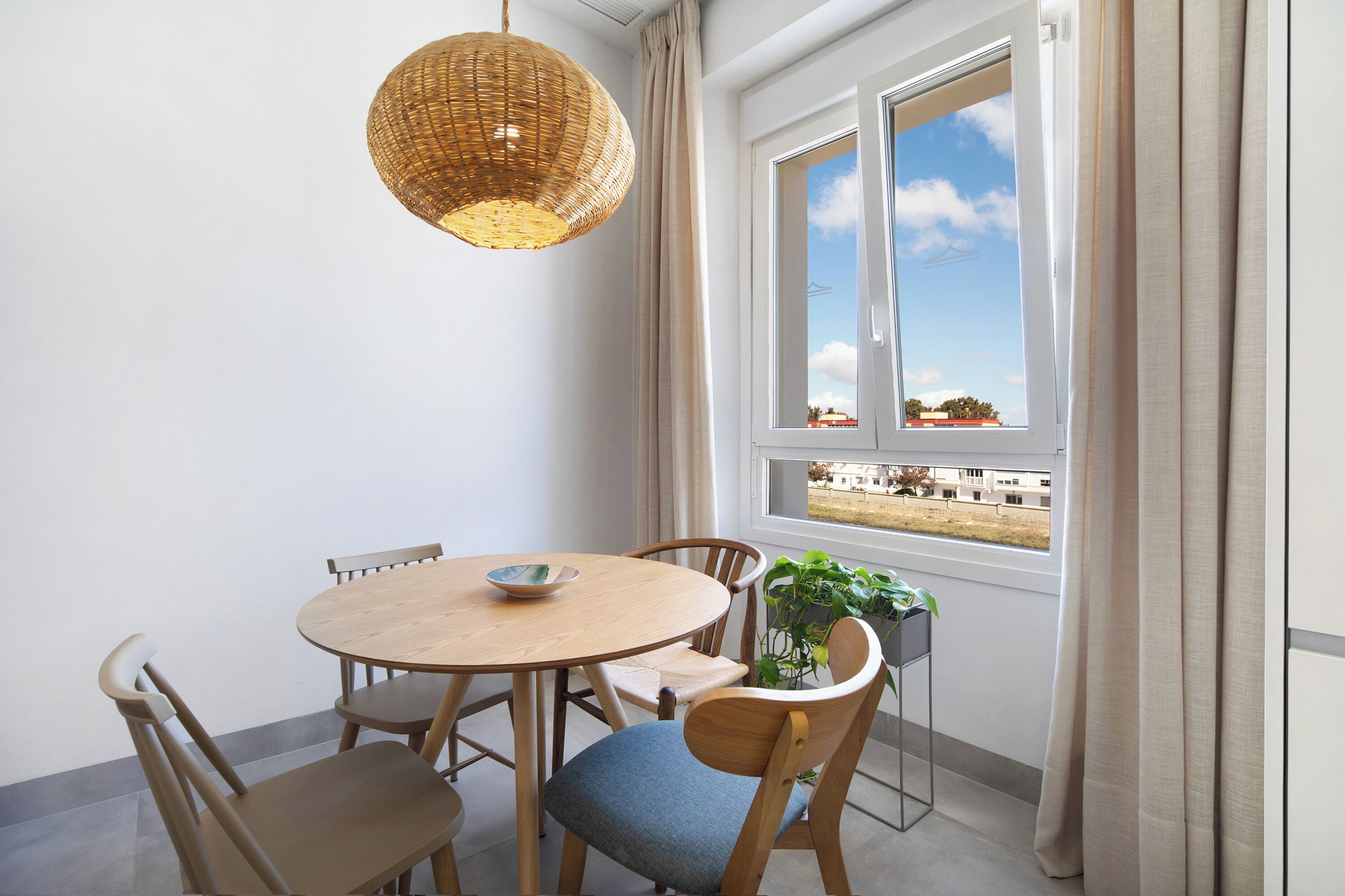 A dining area with a round wooden table, four chairs, a hanging wicker light fixture, a potted plant, and a window with curtains.