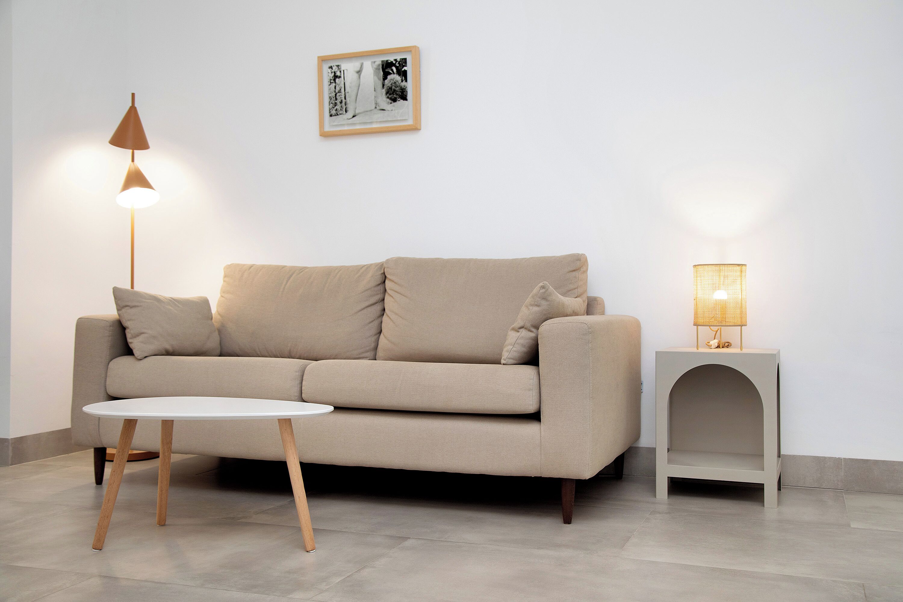 A neutral-toned living room with a beige sofa, a round white coffee table, a floor lamp, and a side table with a lamp.